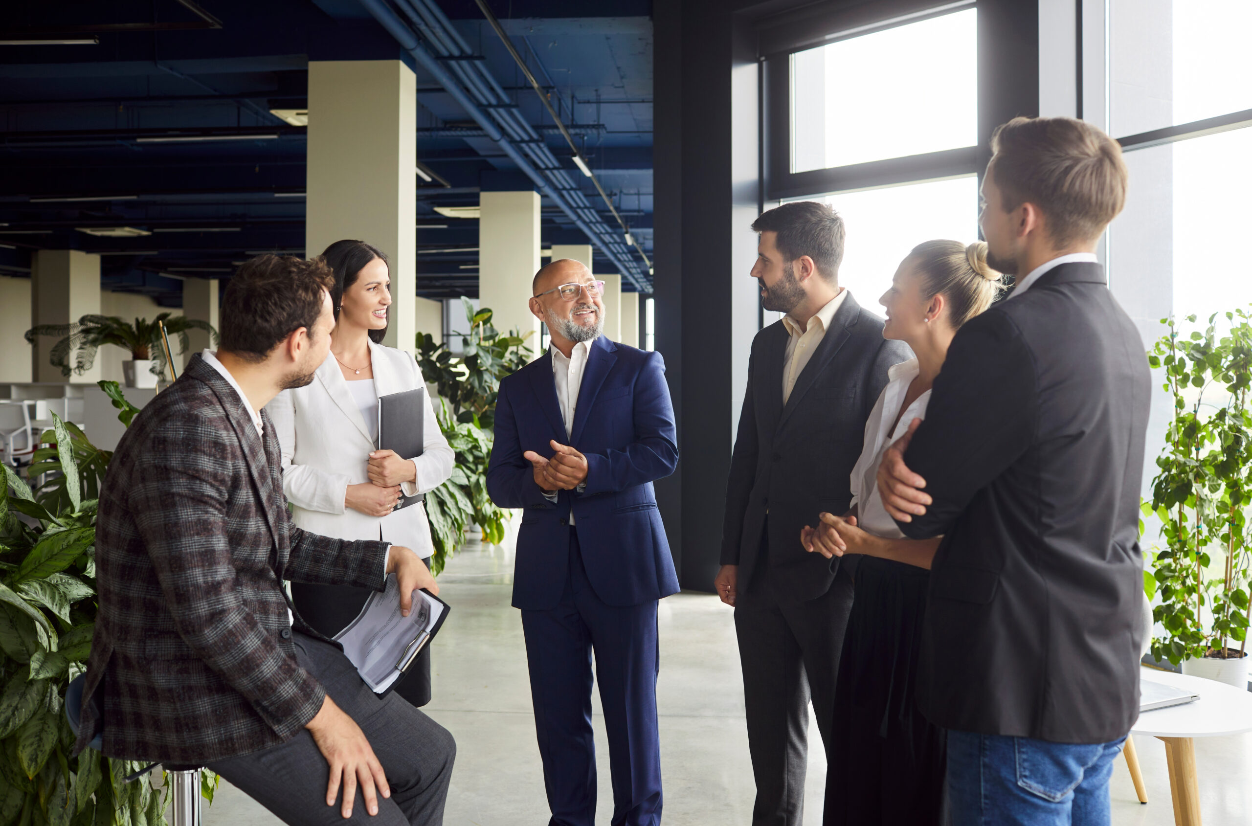 Business meeting of colleagues team and senior corporate leader, standing in modern open space office. People talk with smiles, listen and share ideas and plans of leadership and teamwork